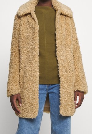Personne portant un manteau en fausse fourrure bouclée beige sur un pull vert olive et un jean bleu sur un fond blanc uni.
