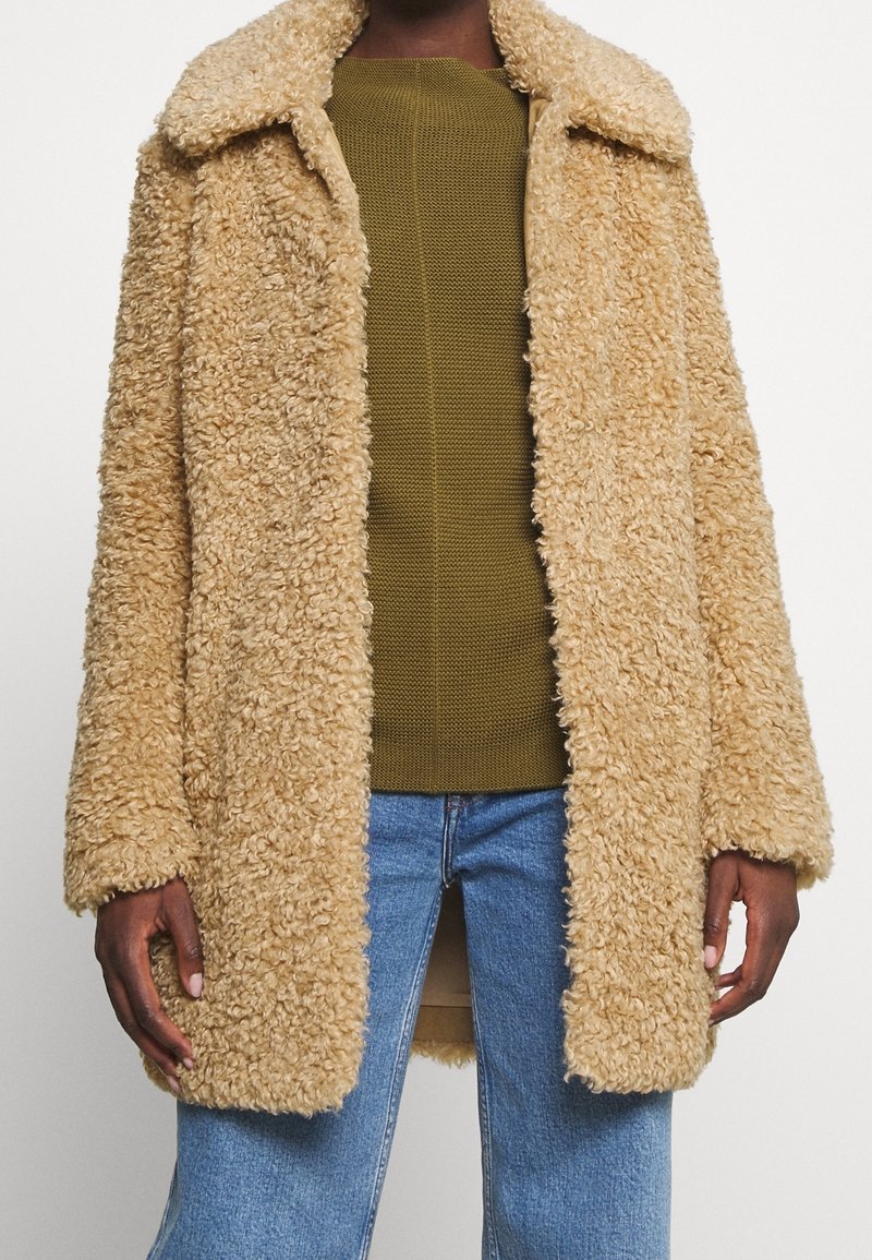 Personne portant un manteau en fausse fourrure bouclée beige sur un pull vert olive et un jean bleu sur un fond blanc uni.