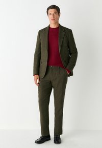 Olivgrön skräddarsydd kostym med en texturerad, ullliknande yta, kombinerad med en burgunderröd rundhalsad tröja och svarta skor. Slim-fit design.