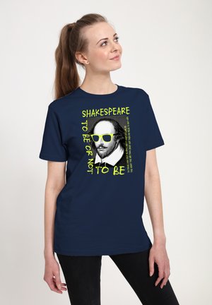 Kvinde iført en marineblå skjorte med en grafik af Shakespeare, der bærer gule solbriller, og teksten "Shakespeare to be or not to be".