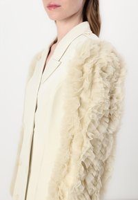 Blazer créme avec des manches en tulle beige à volants. Présente un col à revers et trois boutons à l'avant, créant une texture superposée.