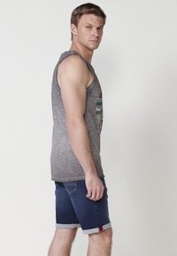 Siva brezrokavnik s grafičnim dizajnom, v kombinaciji z modrimi denim kratkimi hlačami z zavihanimi robovi in majhnim rdečim poudarkom na robu.