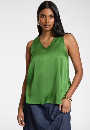 Donna con capelli lunghi e neri, che indossa un top di raso verde senza maniche, pantaloni blu, orecchini dorati e un braccialetto d'oro, in posa su uno sfondo semplice.