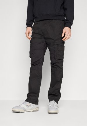 Mann in schwarzen Cargo-Hosen und hellgrauen Sneakers, der vor einem schlichten hellgrauen Hintergrund steht.