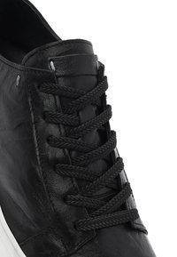 Zwarte leren sneaker met dikke zwarte gevlochten veters, gestrikt door zes paren veteroogjes, op een witte achtergrond.