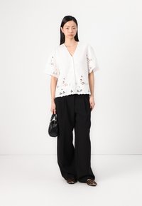 White embroidered blouse with short, flared sleeves, featuring a V-neckline and decorative cutouts. Paired with loose black trousers and a small bag.