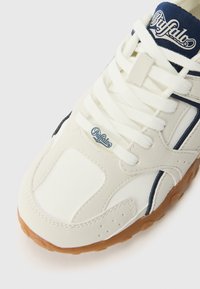 Balti sportbačiai su tekstūruota viršutine dalimi, tamsiai mėlynomis detalėmis ir raišteliais. Yra guminis padas, gumos spalvos, ir logotipas ant liežuvėlio.