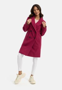 Fuchsia dubbelzijdige jas met grote zwarte knopen, brede revers en zijzakken. Gecombineerd met witte leggings en dikke crèmekleurige sneakers.