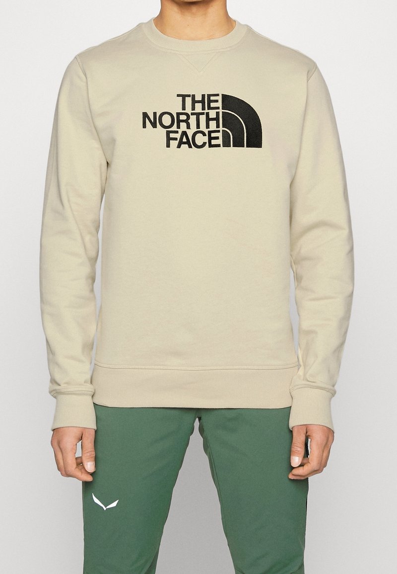 Sudadera beige con un logo negro de "THE NORTH FACE" con un acento curvado, cuello redondo y puños y dobladillo de canalé. Combinada con pantalones verdes.