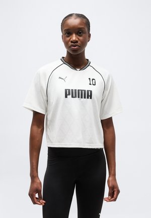 Αθλητική γυναίκα που φοράει λευκό κοντό αθλητικό μπλουζάκι Puma με αριθμό 10 και μαύρο κολάν, όρθια μπροστά σε απλό φόντο.