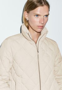 Beige gewatteerde jas met een hoge kraag en zilveren ritsdetails. Textiel met een gewatteerd diamantpatroon, ontworpen voor een getailleerde uitstraling.