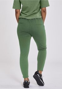 Green leggings made from soft material, featuring a tapered design, elastic waistband, and two back pockets. Worn with black sneakers.