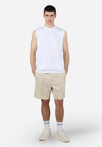 Hvit ärmeløs t-skjorte, beige shorts med frontsting, hvite joggesko og mønstrede ankelsokker. Enkelt, atletisk design med nøytrale farger.