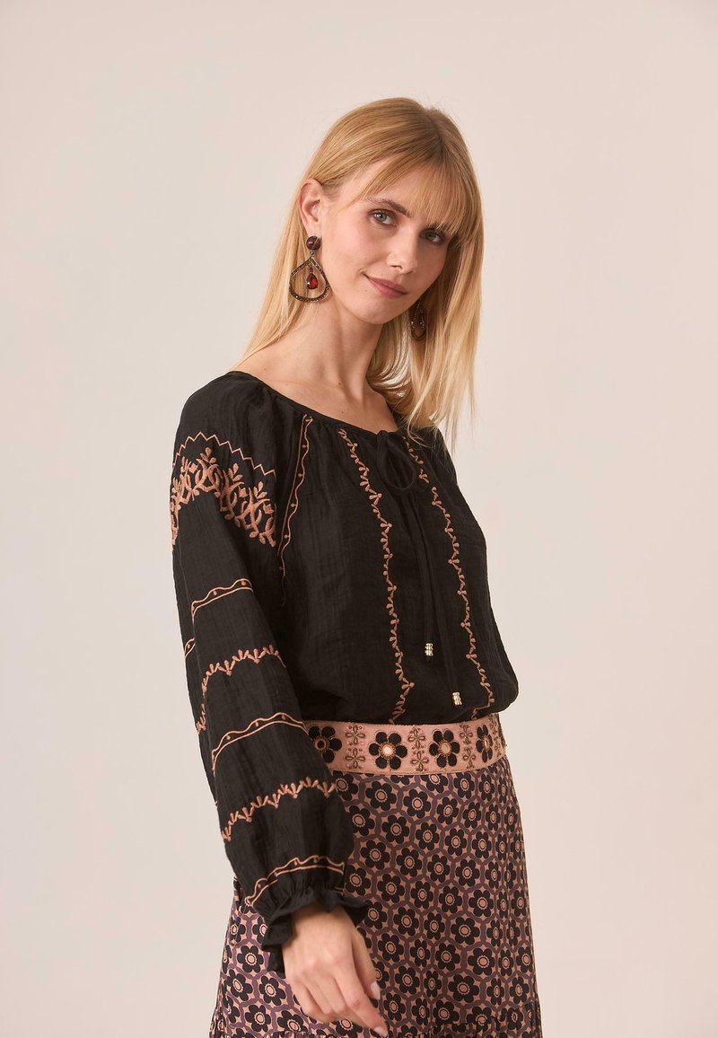 Blusa negra con bordados en rosa y mangas fruncidas, combinada con una falda estampada que presenta diseños florales y una cinturilla rosa.