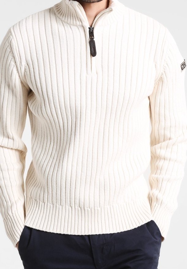Homme portant un pull en tricot côtelé de couleur crème avec un col à fermeture éclair partielle et un pantalon noir, mains dans les poches.