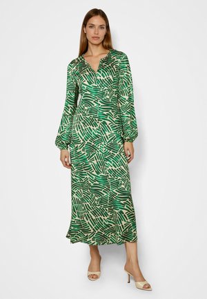 Frau trägt ein langärmeliges Midikleid mit V-Ausschnitt in grün-beigem abstraktem Muster und beige offenen Sandaletten, steht vor einfachem Hintergrund.