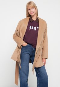 Beige franjitud mantel pehmest kangast, avatud esiosa disain, koos marooni värvi pusaga ja siniste teksadega, näidates juhuslikku stiili.