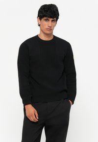 Pulover negru tricotat cu textură rigletă, guler rotund și detalii cu aplicații pe umeri. Asortat cu pantaloni închiși la culoare. Design simplu și curat.