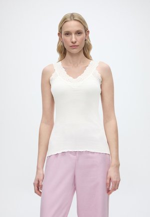 Γυναίκα με μακριά ξανθά μαλλιά που φοράει λευκό αμάνικο τοπ με δαντέλα και ανοιχτό ροζ παντελόνι φαρδύ από κάτω, στέκεται μπροστά σε απλό φόντο.