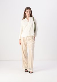 Blusa bianca a maniche lunghe con scollatura a V, abbinata a pantaloni larghi beige chiaro con paillettes e sandali neri, mentre tiene in mano una borsa verde.