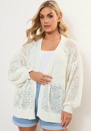 Cardigan bianco lavorato a maglia con motivo a diamante aperto, vestibilità morbida e polsini a coste. Indossato sopra una maglietta bianca e pantaloni shorts di denim.