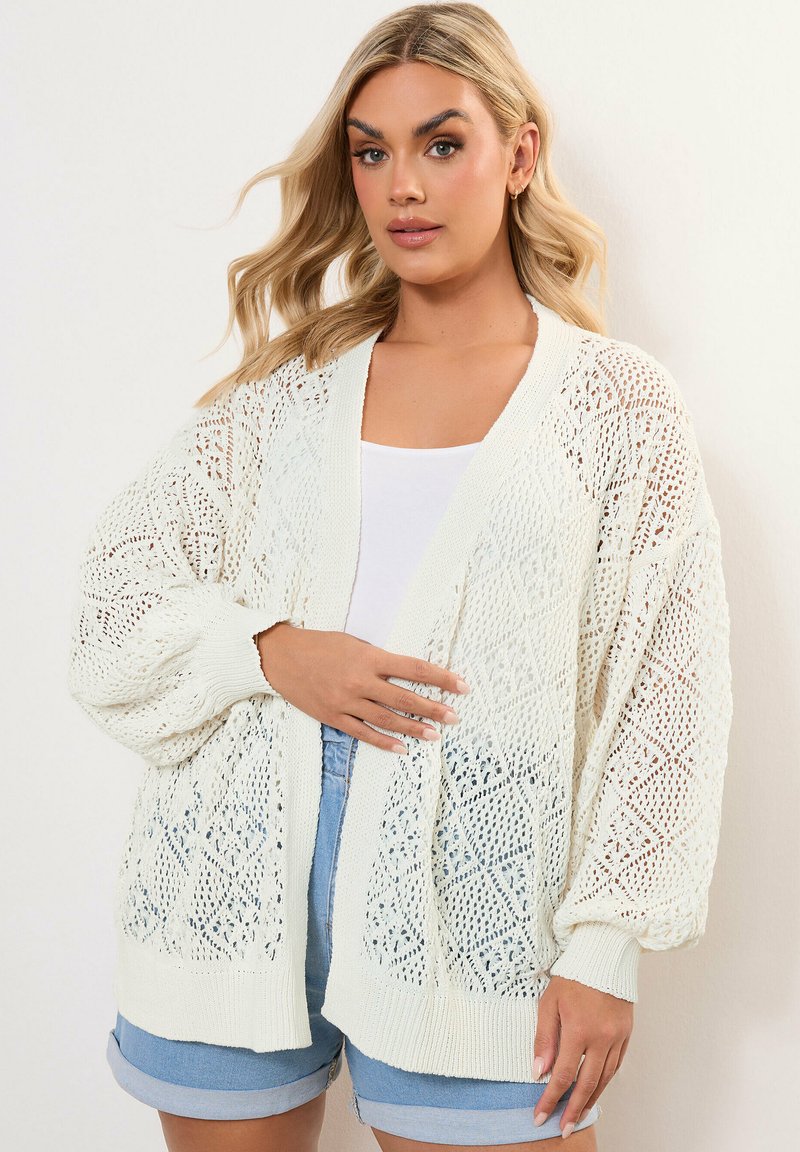 White knitted cardigan with open diamond pattern, loose fit, and ribbed cuffs. Worn over a white top and denim shorts.