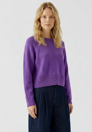 Femme aux cheveux blonds ondulés portant un pull violet à manches longues et un pantalon large sombre, debout devant un fond clair uni.