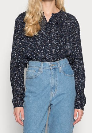 Kobieta ubrana w granatową bluzkę z długim rękawem, ozdobioną małymi białymi i pomarańczowymi kropkami, włożoną do jasnoniebieskich, wysokim stanem spodni jeansowych.