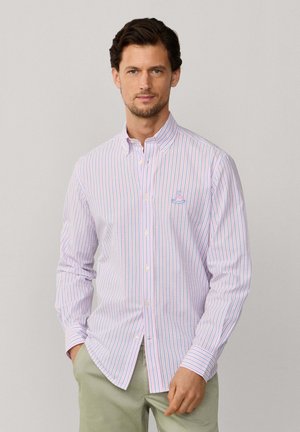 Homme aux cheveux foncés portant une chemise rayée rose et bleue à manches longues boutonnée et un pantalon vert clair, debout devant un fond uni.