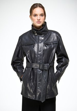 Nő fekete, övvel ellátott bőrkabátban, magas gallérral, cipzáras mellzsebekkel, kezét az oldalzsebekben tartva, fehér háttér előtt áll.