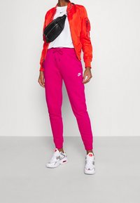 Giubbotto bomber rosso, t-shirt bianca, pantaloni della tuta rosa brillante e scarpe Nike grigie con dettagli. Borsa a tracolla nera.