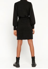 Musta mekko, jossa on kapea siluetti, pitkät puffihihat ja vyö vyötäröllä. Valmistettu teksturoidusta, kimaltelevasta materiaalista; polvipituinen.