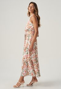 Robe maxi florale à col halter en blanc, avec des motifs orange, rose et vert. Tissu léger avec une silhouette fluide.