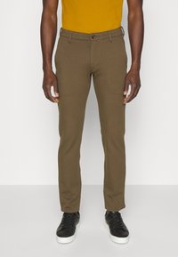Man met slim-fit olijfgroene broek, een mosterdkleurig shirt in zijn broek gestoken en zwarte sneakers met witte zolen, staand tegen een effen achtergrond.