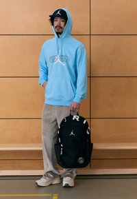 Világoskék kapucnis pulóver logóval, bézs kordbársony nadrág, szürke cipő. Fekete hátizsák fehér felirattal és logóval. Egyszerű, funkcionális dizájn.