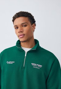 Grøn sweatshrt med lynlås og broderet tekst i pink. Casual design med høj krave og tekstureret stof.