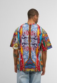 Mann mit kurzem Haar und Tattoo trägt ein übergroßes, mehrfarbig gemustertes Hemd und hellblaue Jeans, blickt von hinten gegen einen schlichten grauen Hintergrund.