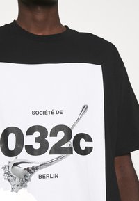 T-shirt noir à manches courtes avec un grand motif blanc sur le devant affichant "SOCIÉTÉ DE 032c BERLIN" et une illustration d'une cuillère métallique.