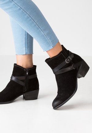 Botas negras de ante hasta el tobillo con tacones bajos de bloque, cremallera lateral y correa decorativa con hebilla, usadas con vaqueros cortos azul claro.