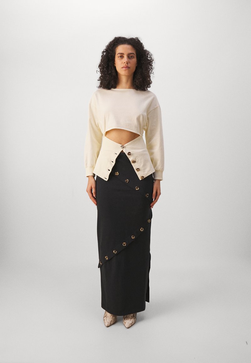 Sweatshirt court crème avec détail en découpes, associé à une jupe noire comportant des accents de boutons diagonaux et une fente sur le côté. Chaussures en peau de serpent texturées.