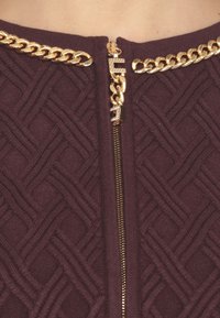 Maglione bordeaux con texture e motivo geometrico, dotato di un accento con catena dorata vicino al colletto e di una cerniera dorata visibile.