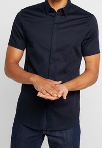Námořnická košile s krátkým rukávem a límcem na knoflíky, vyrobená z hladké tkaniny. Obsahuje decentní logo na hrudi a rovný lem.