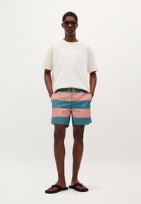 Shorts pour hommes avec des rayures horizontales roses et teal, dotés d'une taille à cordon de serrage et de petits motifs répétitifs. Portés avec un t-shirt blanc et des tongs.