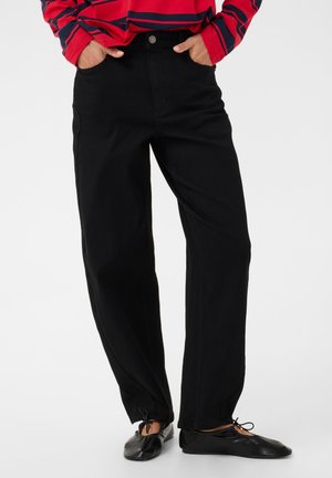 Person trägt schwarze, gerade geschnittene Hose, schwarze Ballerinas und ein rotes Langarmshirt mit marineblauen Streifen, Hände in den Taschen.