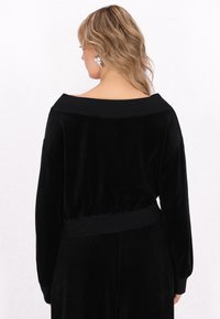 Tuta in velluto nero con una ampia scollatura off-the-shoulder, maniche lunghe e vita aderente. Texture liscia con una silhouette rilassata.