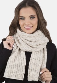 Beige pletená šála s texturovaným vzorem, volně ovázaná kolem krku, s třásněmi na koncích a jemným, teplým vzhledem.