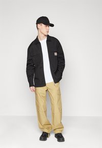 Sort lærredjakke med to brystlommer, hvid t-shirt under, beige cargo bukser og sorte sneakers, sat mod en ensfarvet baggrund.