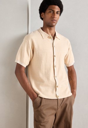 Short-sleeved, light beige shirt with a textured pattern, collared neckline, and button closure, paired with brown trousers.