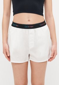 Vita bomullsshorts med svart elastisk midjeband som har en flerfärgad "Calvin Klein"-logotyp. Lätt och slät textur.
