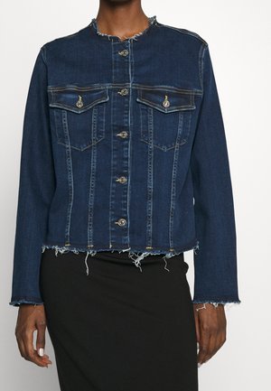 Chaqueta de mezclilla azul oscuro con dobladillo y puños deshilachados, cierre de botones y dos bolsillos en el pecho, usada con falda negra.